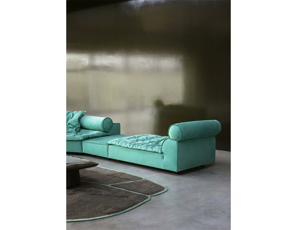 A low, angular sofa in bright turquoise upholstery with soft, voluminous cushions designed for lounging, complemented by minimalist accessories and a dark glass table that add contrast to the vibrant composition.