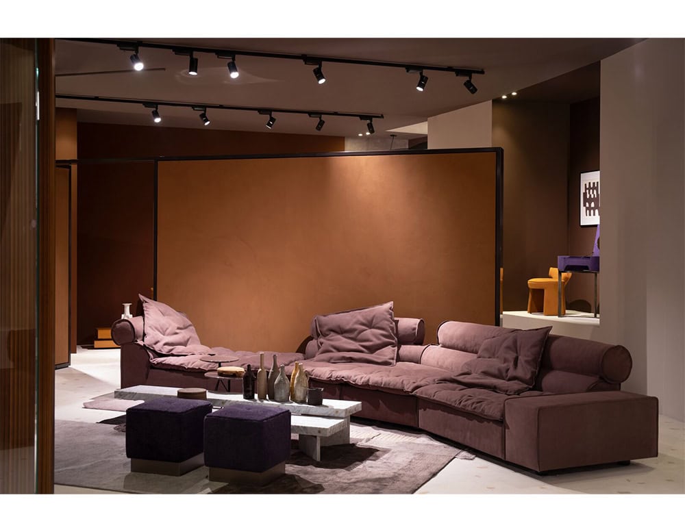 An elegant sectional sofa in velvety plum upholstery featuring generous seating and cylindrical backrests, complemented by minimalist tables and rich earthy hues that enhance its refined, cozy presence.