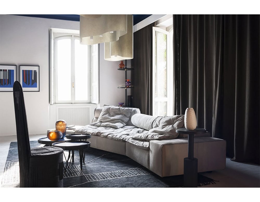 A curved gray sofa with plush cushioning and a low modern silhouette anchors the room’s sophisticated palette while paired with sculptural tables and warm amber glass vases for a refined, inviting ambiance.