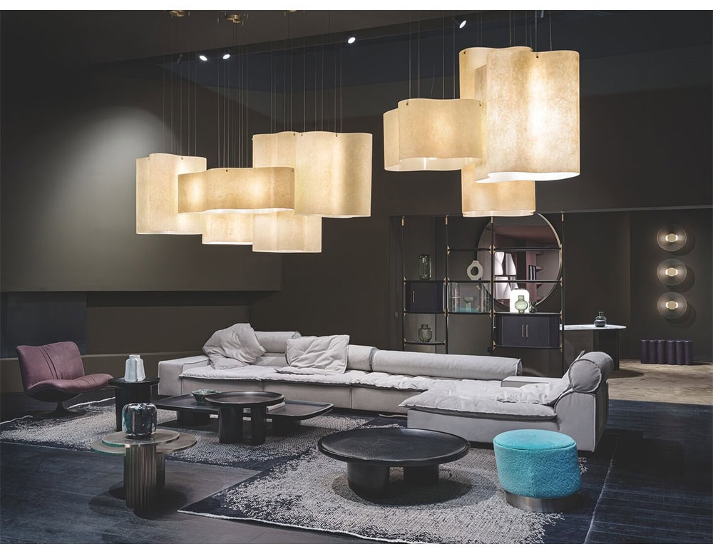 Set in a refined interior, this modular sofa in soft gray tones features plush cushions and generous proportions complemented by dark circular tables and a turquoise pouf that add subtle contrast beneath sculptural pendant lighting.