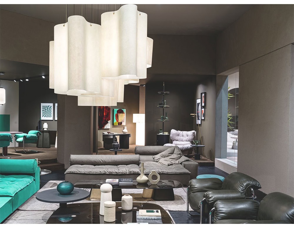 A contemporary sectional sofa in soft gray anchors the space with its relaxed silhouette and layered cushions paired with a teal accent sofa, dark leather chairs, and sculptural tables beneath a bold pendant light installation.