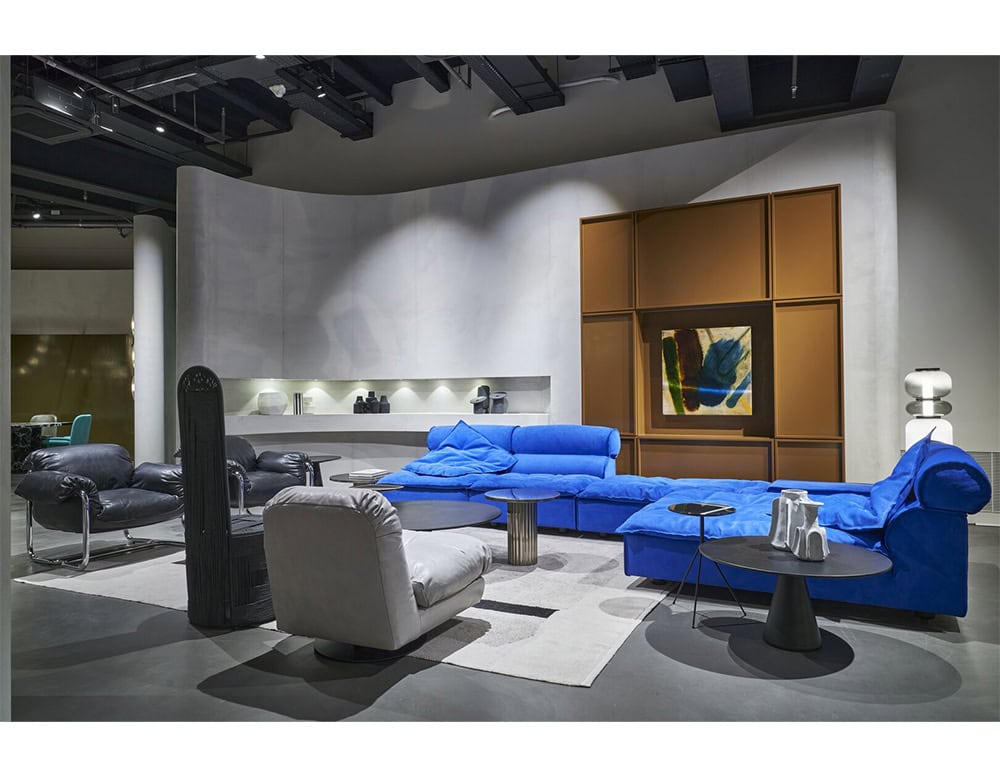 A striking blue sectional sofa anchors the room with bold color and soft contours, its generous cushions creating an inviting centerpiece among sculptural tables and modern armchairs.