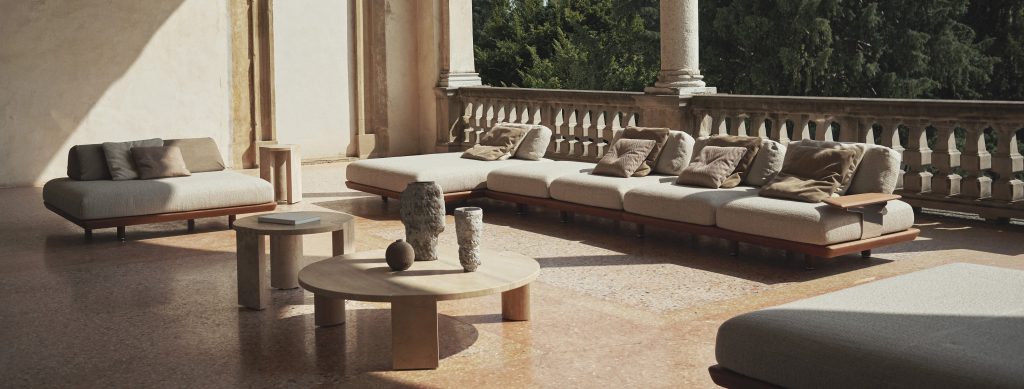 An elegant outdoor terrace featuring modular sofas with soft neutral cushions, complemented by round wooden tables and sculptural decor, set against a sunlit stone backdrop and lush green views. An elegant outdoor terrace featuring modular sofas with soft neutral cushions, complemented by round wooden tables and sculptural decor, set against a sunlit stone backdrop and lush green views.