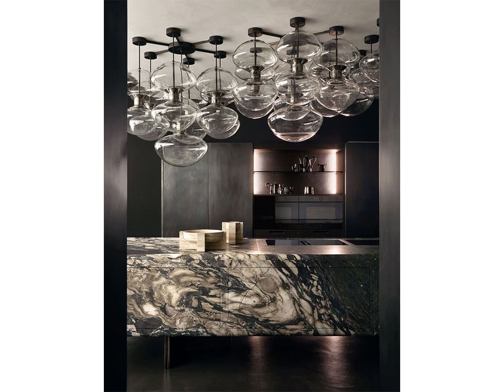 Rounded glass ceiling lights float in an elegant arrangement, diffusing warm reflections across a sophisticated kitchen with a richly veined marble island and dark minimalist cabinetry below.