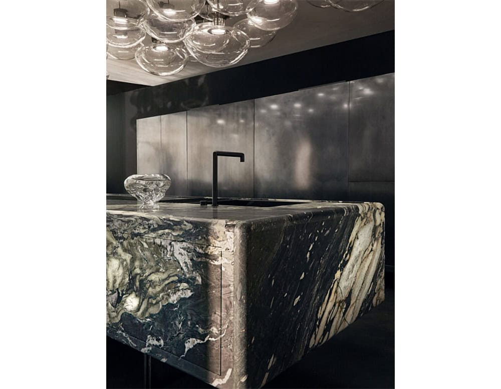 A cluster of seven clear glass ceiling lights hangs gracefully above a sleek kitchen, their rounded forms reflecting soft light over a sculptural marble island and dark, minimalist cabinetry below.