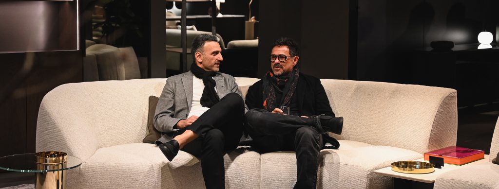 Two people sit comfortably on a light sofa in a modern, softly lit interior, engaged in a relaxed conversation with warm decor elements around them.