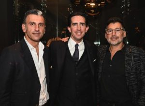 A trio of well dressed guests share a poised moment together, standing confidently with relaxed smiles that reflect the welcoming and sophisticated atmosphere of the evening.