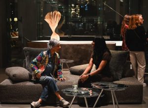 Two guests settle into a cozy conversation on a sculptural sofa, surrounded by soft lighting and striking decor. The relaxed moment highlights the inviting energy of the showroom gathering.