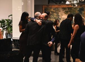 A heartfelt embrace becomes the center of the room as guests gather around in conversation. The atmosphere feels joyful and relaxed, capturing a genuine moment of connection in the elegant showroom setting.