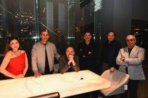 A group poses around a glowing table, each person showing a relaxed and confident expression. Warm light highlights their presence, creating a refined and welcoming atmosphere in the modern setting.
