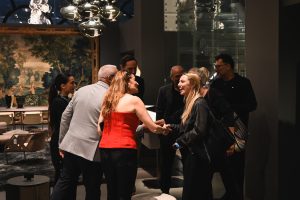 A warm handshake takes place between two women as guests mingle around them. Smiles, lively conversation, and elegant decor create a welcoming and sophisticated atmosphere in the room.