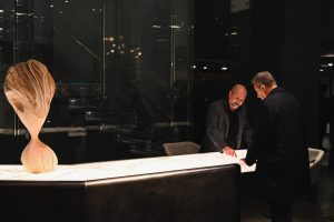 Two men share a warm, engaged conversation while examining a material sample on a long illuminated table. A sculptural wooden art piece adds elegance to the modern, dimly lit interior.