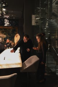 People gather around a glowing table in a modern interior, chatting and observing the surface as warm lighting and reflective decor create an inviting event atmosphere.