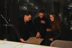 Three people gather around a glowing table in a modern interior, examining a material sample while holding drinks, creating a focused yet relaxed event atmosphere.