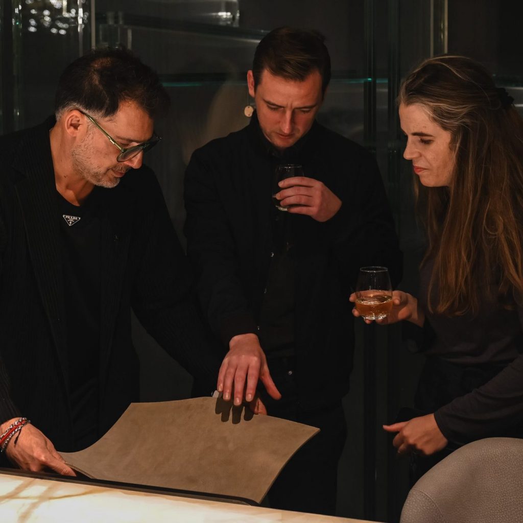 They examine a material sample together, each focused and engaged. Soft lighting highlights their expressions while their drinks rest in hand, creating a calm and thoughtful moment in the design setting.