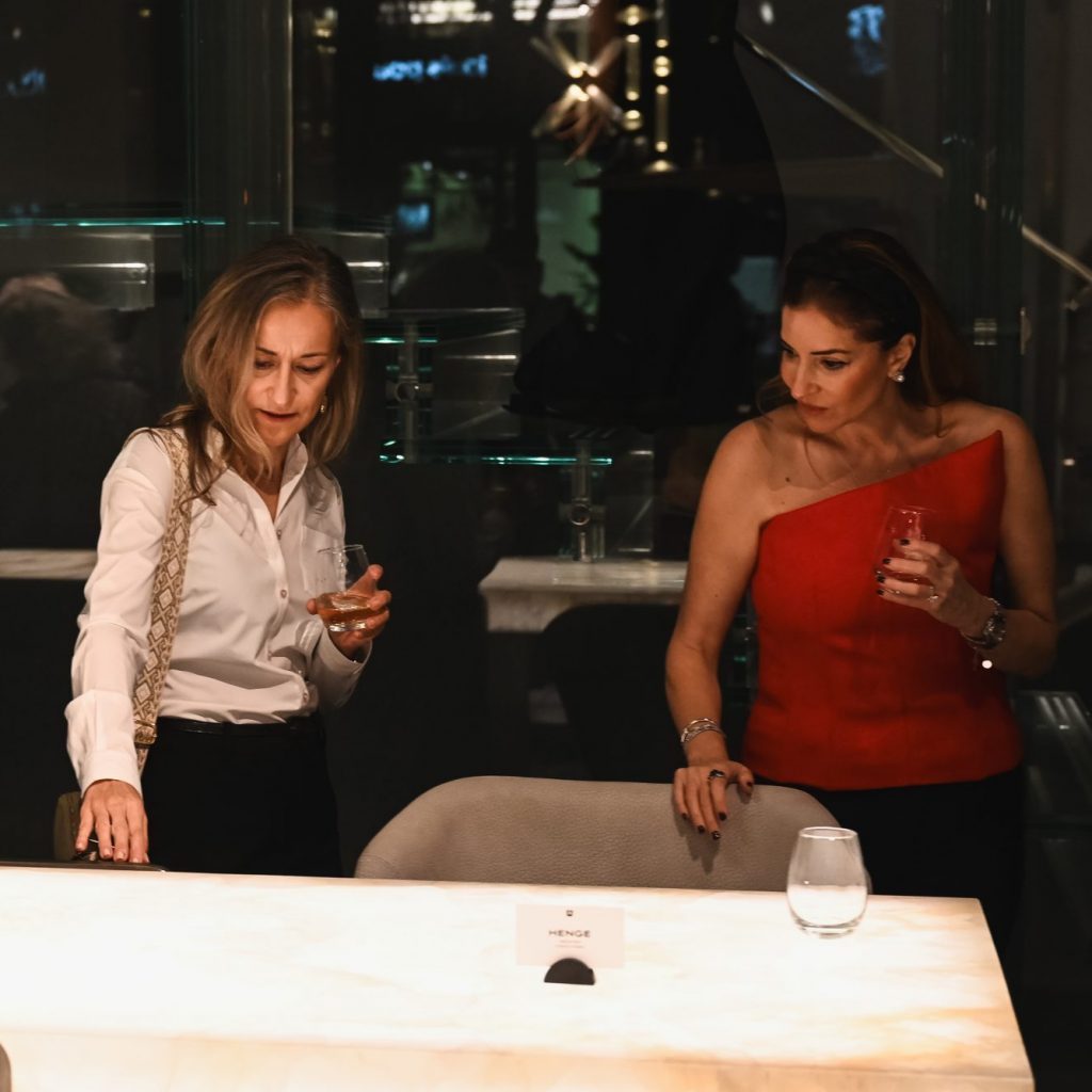 They stand by the glowing table, drinks in hand, observing its surface with focused curiosity. The warm light highlights their attentive expressions and creates an elegant, intimate moment within the design setting.