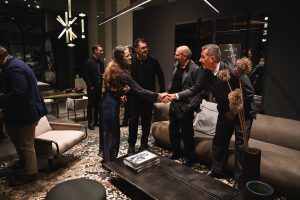 People greet each other warmly in a modern interior, shaking hands and smiling while gathered around contemporary seating and artistic lighting that enhance the welcoming event atmosphere.