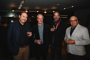Four people stand together at an event, holding drinks and smiling in a warmly lit modern interior with a relaxed and welcoming atmosphere.