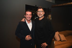 Two people pose together at an event in a modern interior, smiling warmly with one holding a drink and comfortable seating visible in the background.