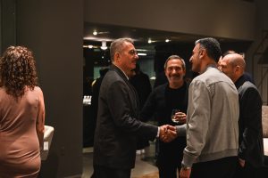People greet each other warmly at an event in a modern interior, shaking hands and sharing smiles while others converse nearby in a relaxed atmosphere.
