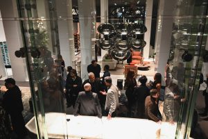 Guests mingle throughout a stylish interior during an event, gathering beneath reflective hanging lights and around modern furniture in a lively and welcoming atmosphere.