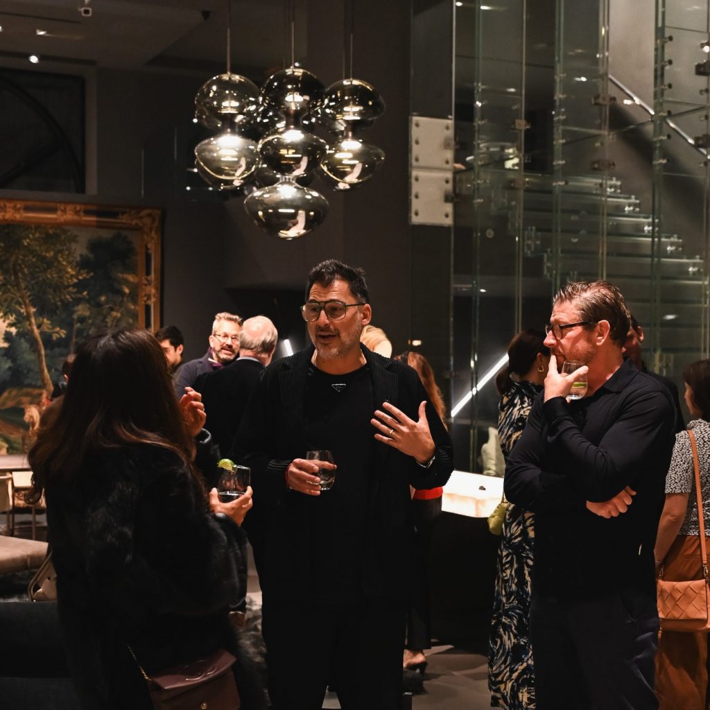 People gathered in a modern interior during an event, engaged in conversation with drinks in hand beneath reflective hanging lights that add a stylish glow to the space.