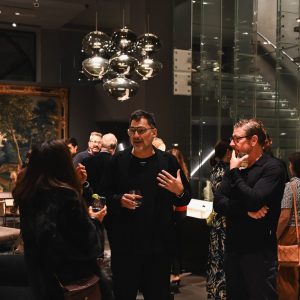 People gathered in a modern interior during an event, engaged in conversation with drinks in hand beneath reflective hanging lights that add a stylish glow to the space.