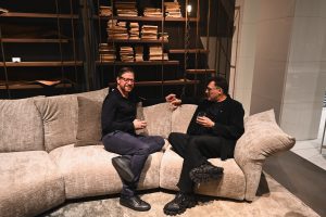 Two people sit comfortably on a plush sofa in a warm modern setting, sharing a relaxed conversation with drinks in hand and shelves of books creating a cozy backdrop.