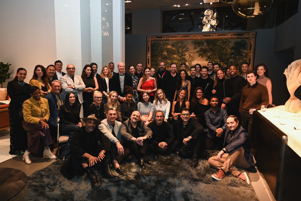 A large group of people gathered in a stylish interior, smiling and posing together on a plush rug and surrounding furniture, creating a warm and festive event atmosphere.