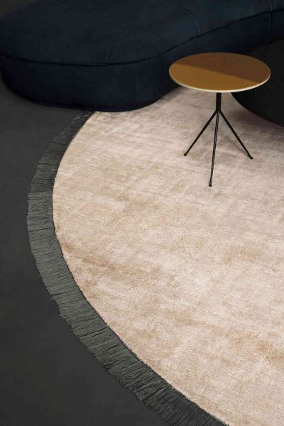 Soft beige Kalahari Rug with a dark fringe border displayed beneath a curved navy sofa and a sculptural round side table. The close up highlights the rugs rich texture and elegant shape within a modern showroom setting.