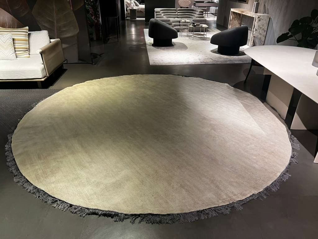 Large round Kalahari rug in a soft neutral tone with textured fringe, displayed in a modern showroom setting where its warm surface contrasts elegantly with the dark flooring and surrounding contemporary furniture.