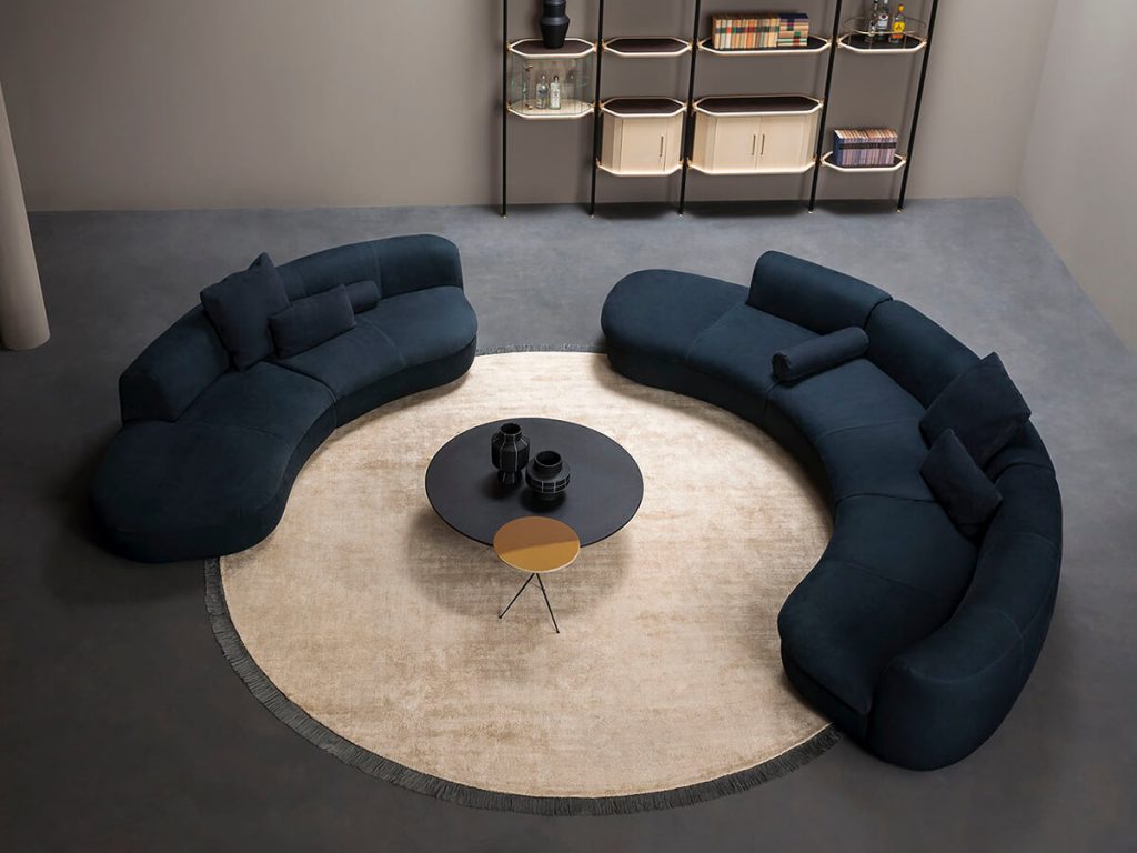Circular beige rug with a plush finish and subtle fringe trim that highlights its shape. It anchors the curved sofas around it, giving the setting a warm and polished look.