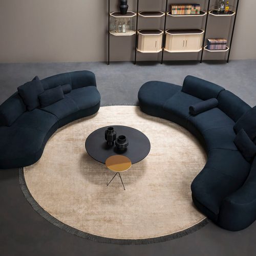 Circular beige rug with a plush finish and subtle fringe trim that highlights its shape. It anchors the curved sofas around it, giving the setting a warm and polished look.