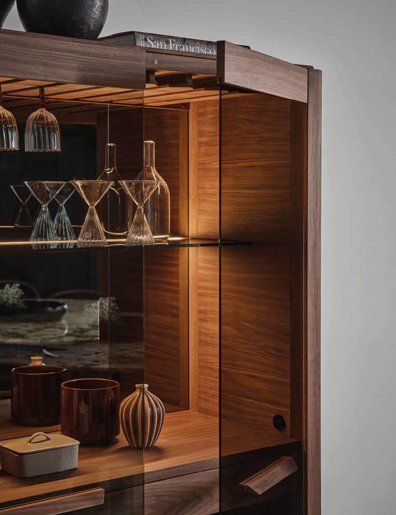 Detail view of the Atlante bar cabinet showing warm walnut wood glass shelving and integrated lighting designed to display bottles and glassware with elegant proportions and refined craftsmanship.