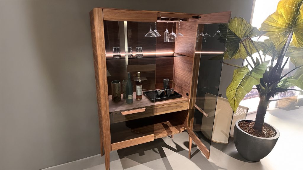 Atlante bar cabinet by Porada shown open with walnut structure glass doors and integrated lighting revealing hanging stemware interior shelves and dedicated storage for bottles and accessories.