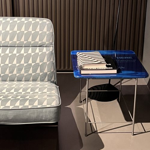 This small accent table features a rich blue glass top with layered tonal variations and subtle graphic details. The glossy surface is supported by a slim silver metal frame, adding a modern touch with a bold pop of color.