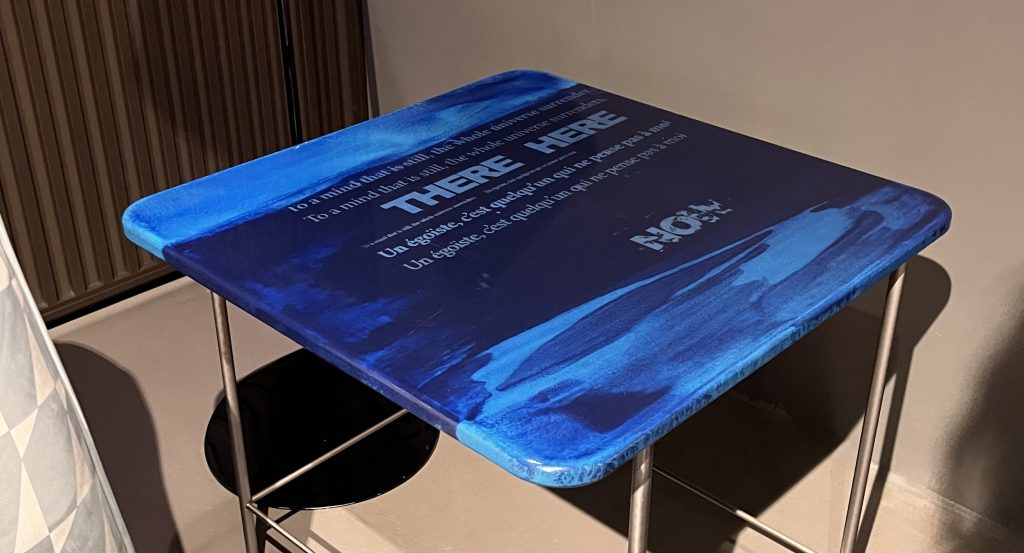 This compact side table features a deep blue glass top with layered tones and subtle graphic text details. The glossy surface rests on a slender metal frame in a light silver finish, creating a modern accent with artistic character.