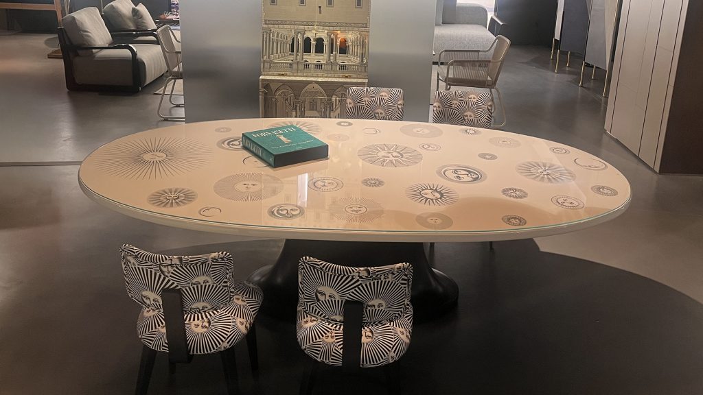 The Soli e Lune table by Fornasetti presents an oval top decorated with iconic sun and moon faces in soft neutral tones creating a refined and playful surface that blends art craftsmanship and functional design.