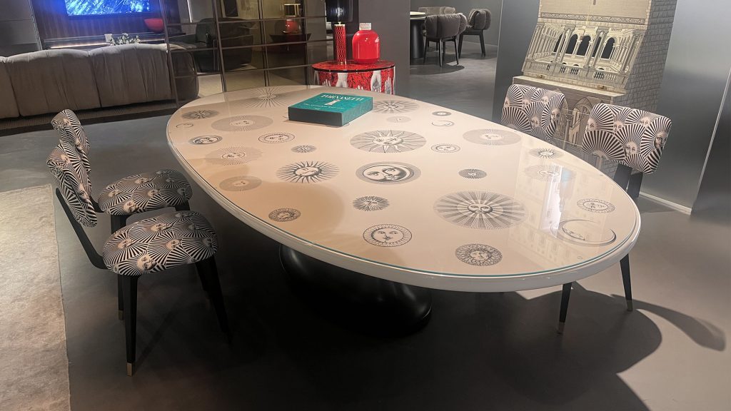 Table Soli e Lune by Fornasetti showcasing an oval tabletop decorated with sun and moon illustrations in soft neutral tones finished with a high gloss surface and supported by a bold central base that blends art and function.