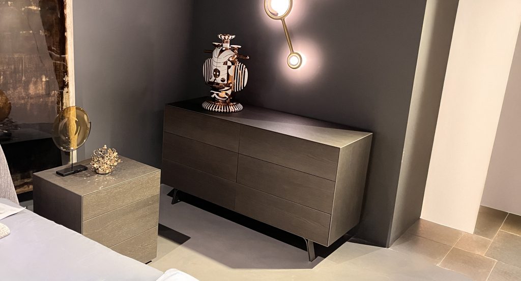 Coordinated dresser and nightstand in deep charcoal-brown wood with a smooth matte finish. Clean, minimalist forms sit on dark legs, paired with warm brass lighting and soft gray surroundings for a refined, contemporary bedroom look.