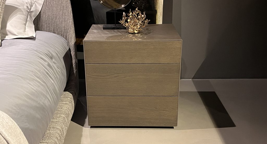 Compact three-drawer nightstand in light taupe wood with a smooth matte finish. Subtle grain and warm neutral tones pair with soft gray textiles, offering a refined, modern bedside accent.