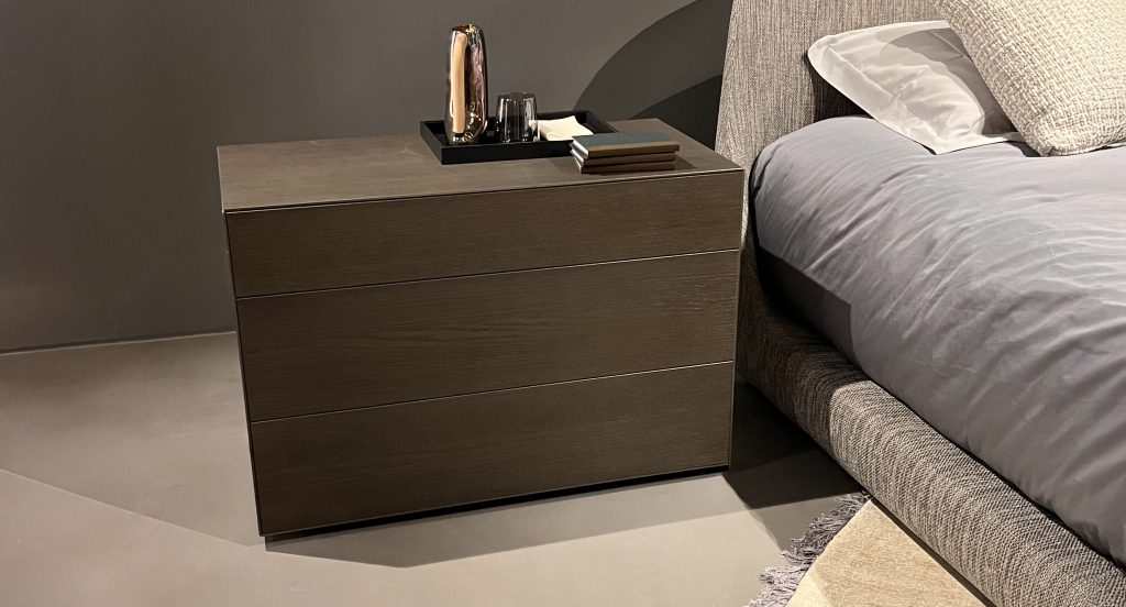 Minimal three-drawer nightstand in warm charcoal wood with a smooth matte finish. Clean lines and low profile pair with soft gray textiles and muted tones, creating a calm, contemporary bedside composition.