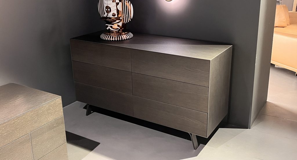 Sleek dresser with clean lines in dark charcoal wood, paired with matte black legs. Set against deep gray walls, warm lighting and a sculptural accent add contrast, highlighting the piece’s modern, refined presence.
