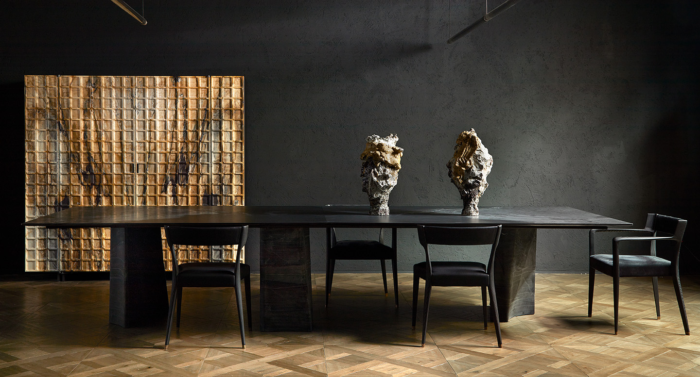 Moody dining scene from the Spiga Milan 2025 display, featuring a dark stone table and black chairs against charcoal walls. Warm bronze and gold accents, sculptural pieces, and wood flooring add depth and contrast.
