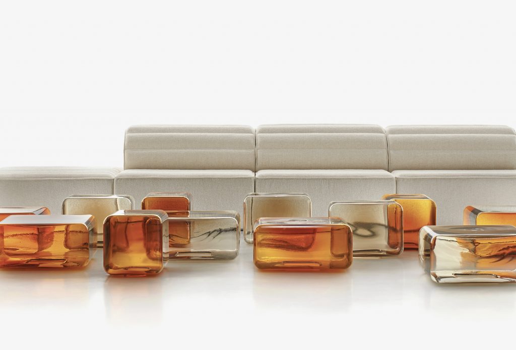 A soft, neutral sofa is paired with a series of translucent amber and clear glass tables, creating a layered, luminous composition. The interplay of warm tones and clean lines adds depth and a refined, contemporary feel.