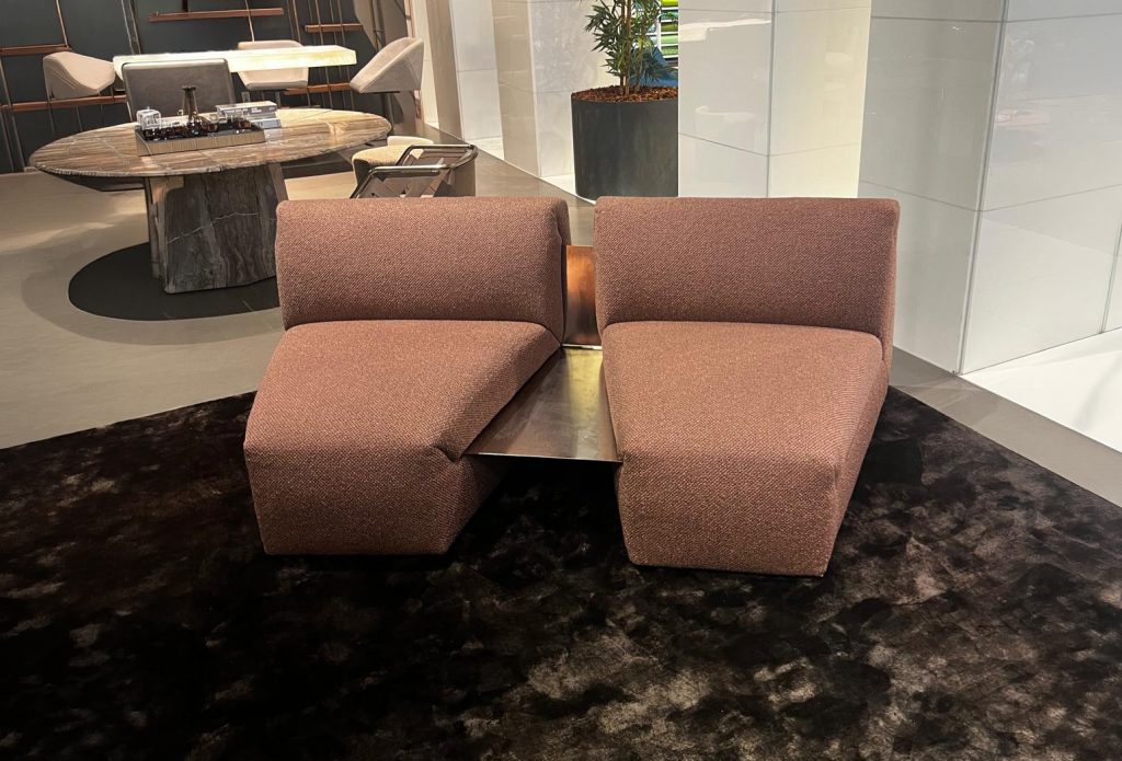 A compact sectional with sculptural geometry and rich textured upholstery. Its angular forms are balanced by a central surface element, creating a functional and visually striking piece suited for modern, intimate lounge settings.