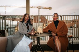 Two elegantly dressed women sit at a small table on a rooftop terrace, smiling against an urban backdrop. Warm tones, soft light, and relaxed posture create a refined yet inviting atmosphere with a contemporary city feel.