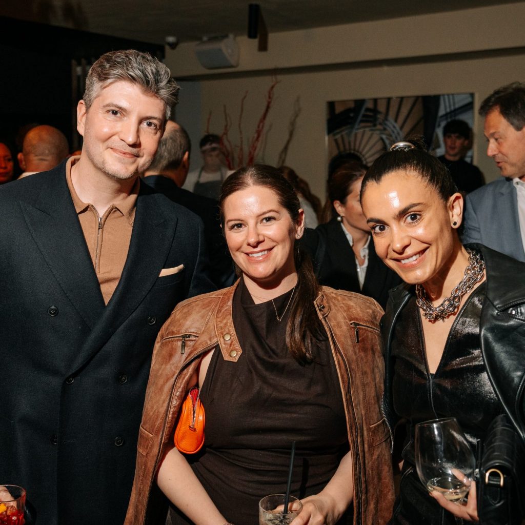 Three guests pose together at a social gathering, smiling in a warmly lit interior. Rich textures and layered outfits add depth, while the relaxed expressions convey an inviting, stylish atmosphere.