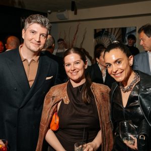 Three guests pose together at a social gathering, smiling in a warmly lit interior. Rich textures and layered outfits add depth, while the relaxed expressions convey an inviting, stylish atmosphere.