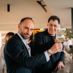 Two guests share a cheerful toast at the bar, raising their glasses with relaxed smiles. The warm indoor setting and casual elegance convey a friendly, celebratory moment during a refined social gathering.